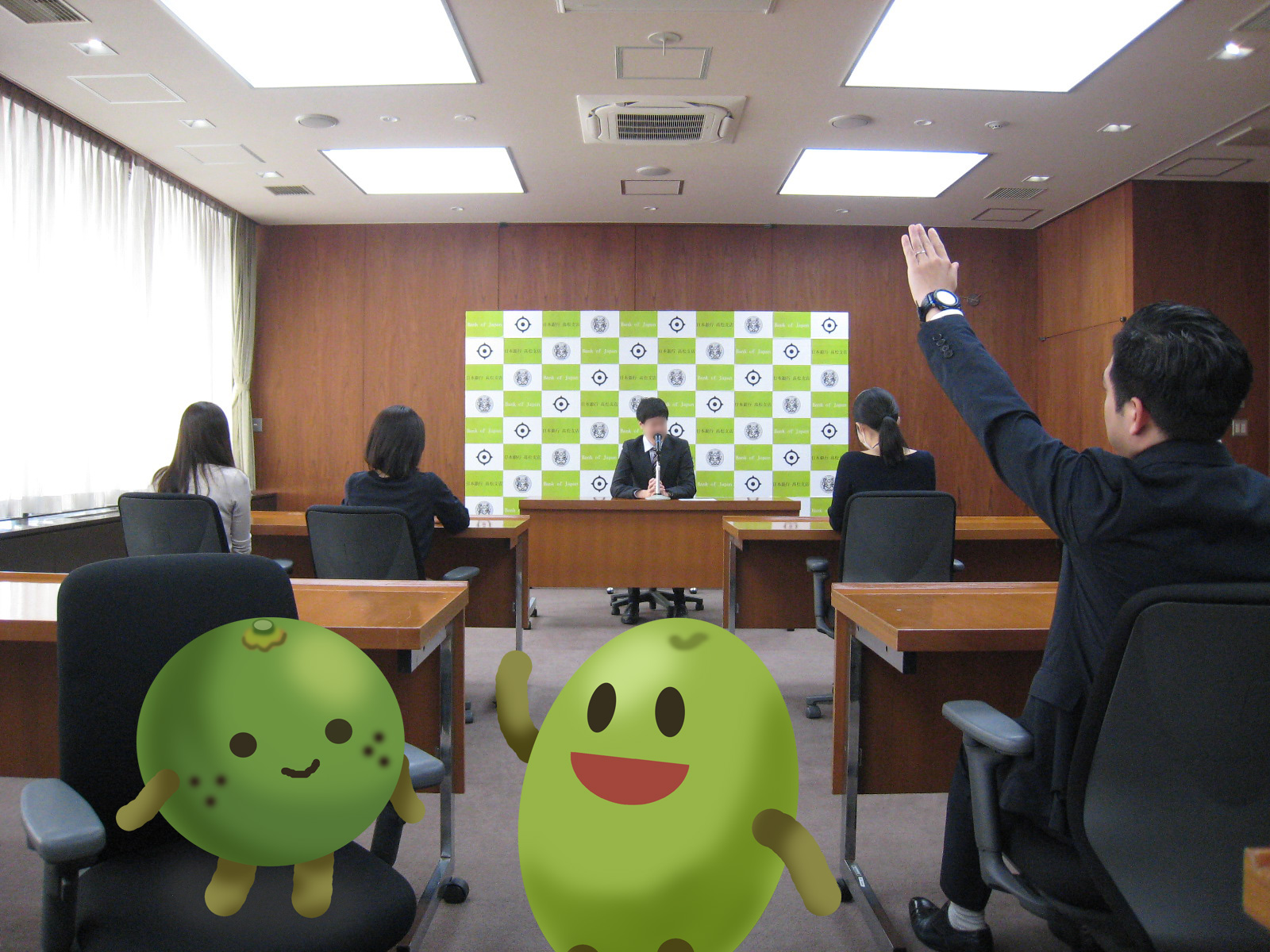 日銀高松支店の会議室で、記者会見を行っている風景を撮影。奥側に会見を実施する職員が着席。手前側には、記者が着席している。記者の１人が手を挙げて質問している